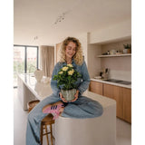 Een vrouw met krullend blond haar, gekleed in spijkerbroek en gestreepte sokken, zit glimlachend op het aanrecht terwijl ze de Kunstplant Gardenia UV 39 cm vasthoudt – een potplant met gele bloemen. Op de achtergrond is een moderne keuken met houten kasten en planken te zien.