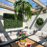 Een zonnig terras met beige zitmeubels met kussens en een houten tafel, versierd met een ananasvaas met roze bloemen en een schaal met aardbeien. Groene wandplanten zorgen voor een weelderige inrichting, aangevuld met de Kunst hangplant varen UV 60 cm in ring zwart als opvallend hangend accent.