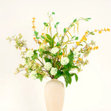 Het Zijden kunstbloemen boeket Golden Meadow bestaat uit groene bladeren, witte hortensiabloemen en gele bloesems in een beige vaas van keramiek - een elegant handgebonden arrangement tegen een effen witte achtergrond.
