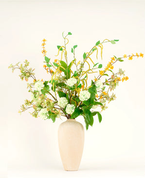 Het Zijden kunstbloemen boeket Golden Meadow bestaat uit groene bladeren, witte hortensiabloemen en gele bloesems in een beige vaas van keramiek - een elegant handgebonden arrangement tegen een effen witte achtergrond.