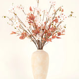 Een crèmekleurige vaas van keramiek bevat takken met delicate lichtroze Zijden kunstbloemen boeket Blushing Whisper en roodbruine bladeren tegen een effen achtergrond - ideaal voor elegante interieurs.