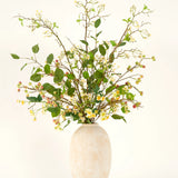 Een hoge vaas van beige keramiek bevat het Zijden kunstbloemen boeket Wild Apple Blossom XL, met groene bladertakken, kleine gele bloemen en roze bessen tegen een lichte achtergrond.