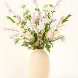 In een vaas met beige structuur staat het Zijden kunstbloemen boeket Lots of Love XL, een handgebonden boeket van roze, witte en blauwe zijden bloemen met groene bladeren en lange stelen tegen een lichte achtergrond.