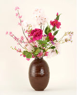 In een vaas met bruine structuur staat het Zijden kunstbloemen boeket Pink Reverie, met roze, magenta en witte kunstbloemen met groene bladeren tegen een effen lichte achtergrond.