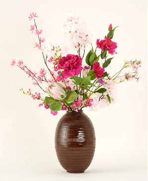 In een vaas met bruine structuur staat het Zijden kunstbloemen boeket Pink Reverie, met roze, magenta en witte kunstbloemen met groene bladeren tegen een effen lichte achtergrond.
