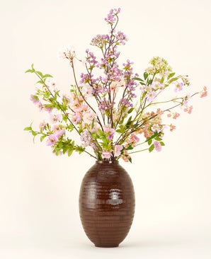 Een bruine vaas van keramiek toont het Zijden kunstbloemen boeket Lilac, een hoog en elegant zijden boeket met roze, paarse en lila bloemen met groene bladeren en dunne takken tegen een effen lichte achtergrond.