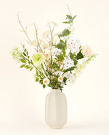 Het Zijden kunstbloemen boeket Claire de Lune heeft delicate witte, lichtgele en groene bloemen in een witte vaas van keramiek, geaccentueerd door weelderig groen tegen een lichte achtergrond.