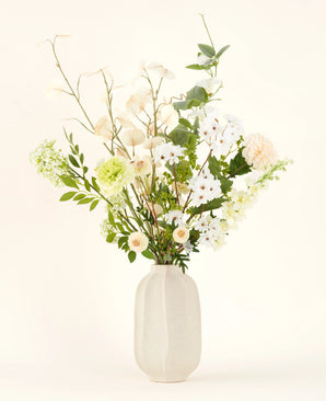 Het Zijden kunstbloemen boeket Claire de Lune heeft delicate witte, lichtgele en groene bloemen in een witte vaas van keramiek, geaccentueerd door weelderig groen tegen een lichte achtergrond.