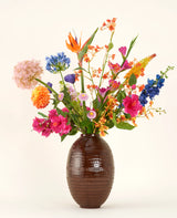 In een vaas met bruine structuur staat het Zijden kunstbloemen boeket Tropical Firework - een arrangement van oranje, roze, paarse, gele en blauwe zijden bloemen met groene bladeren tegen een effen lichte achtergrond.