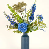 Het Zijden kunstbloemen boeket Mystic Blue Garden bestaat uit blauwe hortensia's, ridderspoor, groene bladeren en gedroogde beige zaadhoofden in een blauwe keramieken vaas - ideaal om je ruimte op te fleuren tegen een lichte achtergrond.