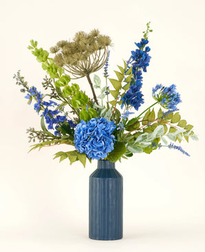 Het Zijden kunstbloemen boeket Mystic Blue Garden bestaat uit blauwe hortensia's, ridderspoor, groene bladeren en gedroogde beige zaadhoofden in een blauwe keramieken vaas - ideaal om je ruimte op te fleuren tegen een lichte achtergrond.