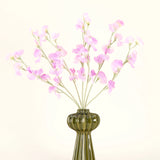 Een groene glazen vaas toont het Zijden kunstbloemen boeket Erwt roze met zachtgroen gebladerte, met verschillende lange stelen en tegen een effen gebroken witte achtergrond.