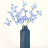 Een blauw geribbelde vaas van keramiek toont het Zijden kunstbloemen boeket Erwt blauw, met zijn delicate lichtblauwe kunstbloemen tegen een effen, lichte achtergrond.