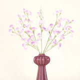 Het Zijden kunstbloemen boeket Erwt lila heeft lange stelen van delicate, lichtpaarse bloemen en groene bladeren in een vaas van mauve keramiek met verticale richels, tegen een effen, gebroken witte achtergrond.