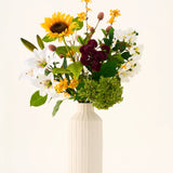 In een crèmekleurige vaas staat het Zijden kunstbloemenboeket ‘Mom’s Garden’, een arrangement van zonnebloemen, lelies, witte en bordeauxrode bloemen en groen blad tegen een effen achtergrond — waardoor het eruitziet als een kunstbloemenboeket dat doet denken aan een zomerse tuinborder.