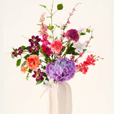 In een witte keramische vaas staat het boeket ‘Rosy Revery’ van Zijden kunstbloemen, een levendig arrangement van roze, paarse, oranje en rode bloemen met groene bladeren en stelen, tegen een lichte achtergrond.