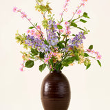 In een donkerbruine vaas staat een boeket van roze, paarse en gele Zijden kunstbloemen, het ‘Sky Kiss’-boeket, met groene bladeren, tegen een effen lichte achtergrond. Deze onderhoudsvrije decoratie zorgt voor blijvende schoonheid in elke ruimte, zonder dat er onderhoud nodig is.