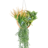 De Kunst hangplant varen UV 30 cm in ring goud heeft verschillende kunstplanten, zoals varens, trapsgewijze wijnranken, stekelig gras en blauwe bloemen, gerangschikt in een gouden ring met een ketting om op te hangen.