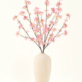 Een hoge vaas van beige keramiek bevat verschillende takken uit het Zijden kunstbloemen boeket Bloemen roze, met delicate roze bloesems en groene bladeren, tegen een effen witte achtergrond.