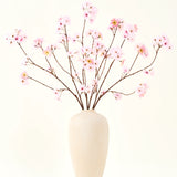 Het Zijden kunstbloemen boeket Kersenbloesem roze 114 cm heeft lange, dunne takken met trossen lichtroze bloemen in een beige vaas van keramiek, die een opvallend contrast vormen tegen de effen witte achtergrond.