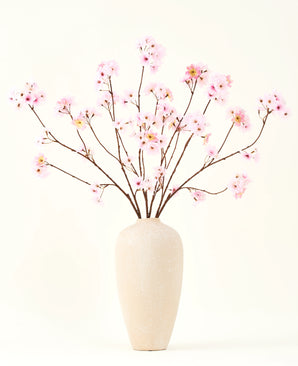 Het Zijden kunstbloemen boeket Kersenbloesem roze 114 cm heeft lange, dunne takken met trossen lichtroze bloemen in een beige vaas van keramiek, die een opvallend contrast vormen tegen de effen witte achtergrond.