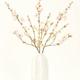 Het Zijden kunstbloemen boeket Kersenbloesem lichtroze 94 cm heeft delicate roze kunstmatige kersenbloesems in een witte keramieken vaas, die elegantie toevoegt tegen een effen lichte achtergrond.