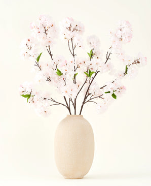 Het Zijden kunstbloemen boeket Kersenbloesem blush 119 cm toont delicate, lichtroze kersenbloesem met groene bladeren in een beige vaas, ideaal als kunstbloemen decoratie tegen een lichte achtergrond.