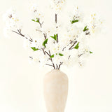 Het Zijden kunstbloemen boeket Kersenbloesem crème 119 cm heeft elegante bruine takken met zacht witte nepbloesem en groene bladeren, weergegeven in een eenvoudige beige vaas tegen een effen lichte achtergrond.