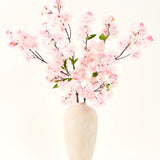 De Zijden kunstbloemen boeket Kersenbloesem roze 119 cm, gerangschikt in een beige keramische vaas met delicate groene bladeren, creëert een sereen interieuraccent tegen een eenvoudige lichte achtergrond.