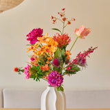 In een witte keramische vaas staat het boeket ‘Magnus di Lambo’ van Zijden kunstbloemen, een kleurrijk arrangement van roze, oranje en gele kunstbloemen met groene bladeren, geplaatst op een houten tafel tegen een beige achtergrond.