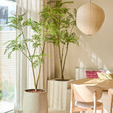 Een lichte eethoek met de Kunstplant Mahonia 210 cm, een grote potplant, een houten tafel en stoelen, een papieren lantaarn en doorschijnende gordijnen die natuurlijk zonlicht binnenlaten. Op een ingebouwde bank liggen roze kussens.