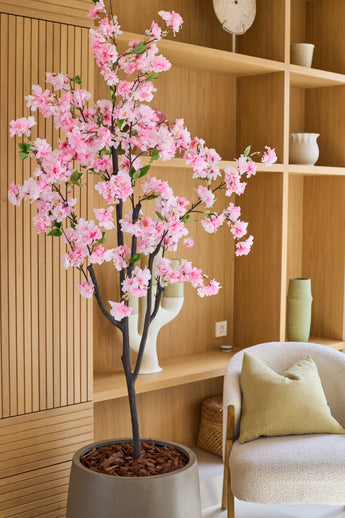 De Kunst Kersenbloesem 150 cm roze, met zijn delicate roze zijden bloesems, staat elegant in een moderne kamer met houten planken, decoratieve voorwerpen, een beige stoel en een groen kussen – en voegt zo de charme van zijden bloemen toe aan je interieur.