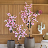 Drie Kunst Kersenbloesem-kunstmatige kersenbloesemboompjes van 180 cm in een roze pot, met roze bloemen, vervaardigd uit hoogwaardige zijden kersenbloesem, staan voor een houten wand met ingebouwde planken, naast een crèmekleurige stoel en decoratieve voorwerpen.