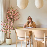 Een vrouw zit glimlachend aan een tafel met een open boek. De kamer is ingericht met lichte houten meubels, papieren lantaarns, zacht natuurlijk licht dat door hoge gordijnen naar binnen valt, en een Kunstplant Kersenbloesem 120 cm roze die een vleugje roze bloesem toevoegt.