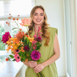 Een glimlachende vrouw met lang blond haar, gekleed in een lichtgroene outfit, staat binnenshuis met in haar handen het Zijden kunstbloemenboeket Magnus di Lambo — een kleurrijk, met de hand gebonden boeket van zijden bloemen, perfect voor wie de voorkeur geeft aan een onderhoudsvrij bloemenarrangement.