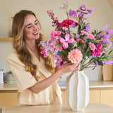 Een glimlachende vrouw met lang blond haar zet een kleurrijk boeket ‘Purple Dawn’ van Zijden kunstbloemen in een witte vaas op het aanrecht.