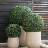 Drie Kunstplant Buxus romeinse stijl UV D40cm kunstheesters in grote beige potten staan op een houten terras, tegen een donkerhouten hek met horizontale latten.