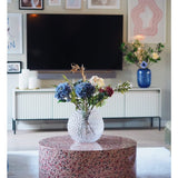 Een moderne woonkamer heeft een terrazzo salontafel met een glazen vaas met bloemen, een tv aan de muur, ingelijste kunst en foto's, een wit dressoir en de Kunst Hortensia Deluxe 57 cm lila als elegant blauw bloemstuk op de achtergrond.