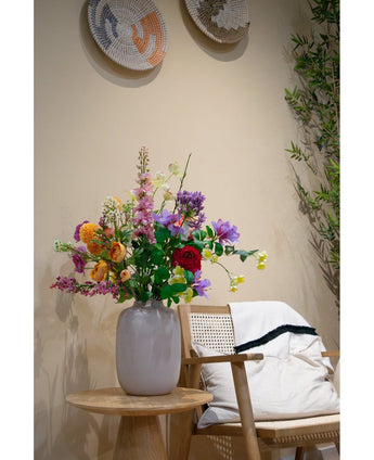 Een Kunst Klaproos Real Touch Paars 65cm staat in een witte vaas op een kleine houten tafel naast een stoel met kussens en een deken. Geweven manden sieren de beige muur en aan de zijkant staat een plant.