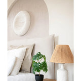 Een keurig opgemaakt bed met wit linnengoed staat naast een houten nachtkastje met daarop de Kunst Monstera 40 cm in sierpot zwart en een witte lamp met een geweven kap. Aan de muur hangt een zachte boog en een rond kunstwerk met structuur.