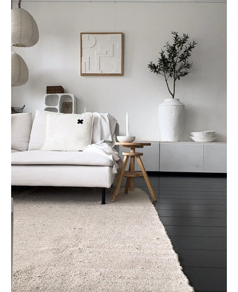Een minimalistische woonkamer met een lichtgekleurde bank, neutraal vloerkleed, wit dressoir, Kunst Olijfboom 120 cm als decoratief element, abstracte muurkunst en houten krukjes-alles in zachte Mediterrane sfeer met neutrale tinten.