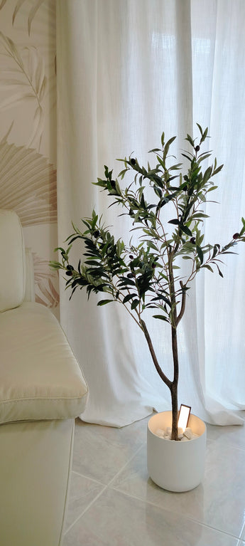 Een Kunst Olijfboom 120 cm met groene bladeren staat op een tegelvloer naast een crèmekleurige bank en geeft de kamer een Mediterrane sfeer, voor witte gordijnen en een muur met subtiel patroon.
