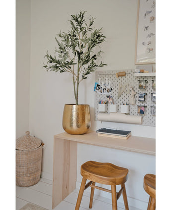 Een moderne werkplek met een houten bureau, twee krukjes, een grote gouden plantenbak met de Kunst Olijfboom 120 cm voor een mediterrane flair, een wasbord met kantoorbenodigdheden en een rieten mand op de vloer als decoratief detail.