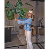 Een lachende vrouw met hoed, blauwe blouse en witte broek staat binnen en houdt een Kunst hangplant Monstera Deluxe 60 cm met grote groene bladeren vast, omringd door modern decor voor een tropische sfeer met decoratieve planten.