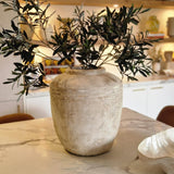 Een rustieke beige vaas met Kunst olijfblad 90 cm brandvertragend staat op een marmeren aanrecht in een moderne, warm verlichte keuken, met planken en mediterrane decoratie op de achtergrond.