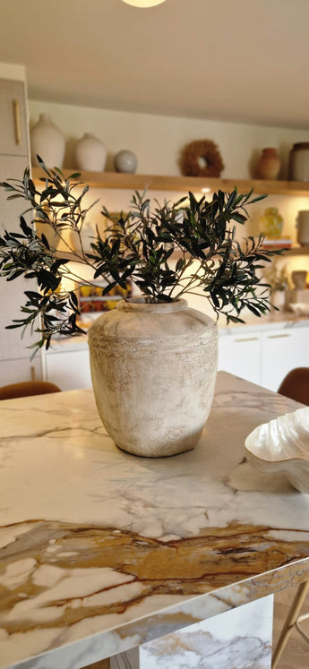 Een rustieke beige vaas met Kunst olijfblad 90 cm brandvertragend staat op een marmeren aanrecht in een moderne, warm verlichte keuken, met planken en mediterrane decoratie op de achtergrond.