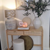 Een houten consoletafel met twee laden bevat een lamp, een grote witte vaas met kunst olijfblad 90 cm brandvertragend, een kleinere vaas, boeken en decoratieve stenen. Mediterrane accenten en ingelijste abstracte kunst maken de look hierboven compleet. Onderaan staan een kruk en een rond dienblad.