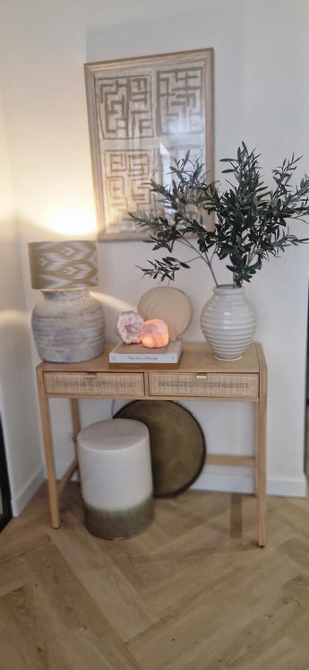Een houten consoletafel met twee laden bevat een lamp, een grote witte vaas met kunst olijfblad 90 cm brandvertragend, een kleinere vaas, boeken en decoratieve stenen. Mediterrane accenten en ingelijste abstracte kunst maken de look hierboven compleet. Onderaan staan een kruk en een rond dienblad.