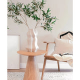 In een moderne, minimalistische leefruimte met mediterrane inrichting staat een gebeeldhouwde witte vaas met het Kunst olijfblad 90 cm brandvertragend op een ronde houten tafel, naast een lichte houten stoel met witte en zwarte kussens.