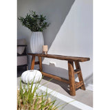Een rustieke houten bank rust naast een witte muur, naast een grote vaas met het Kunst olijfblad 90 cm brandvertragend. Een rond wit kussen en een beige kaars voegen warmte toe en doen denken aan een mediterrane inrichting; gras verschijnt op de voorgrond.